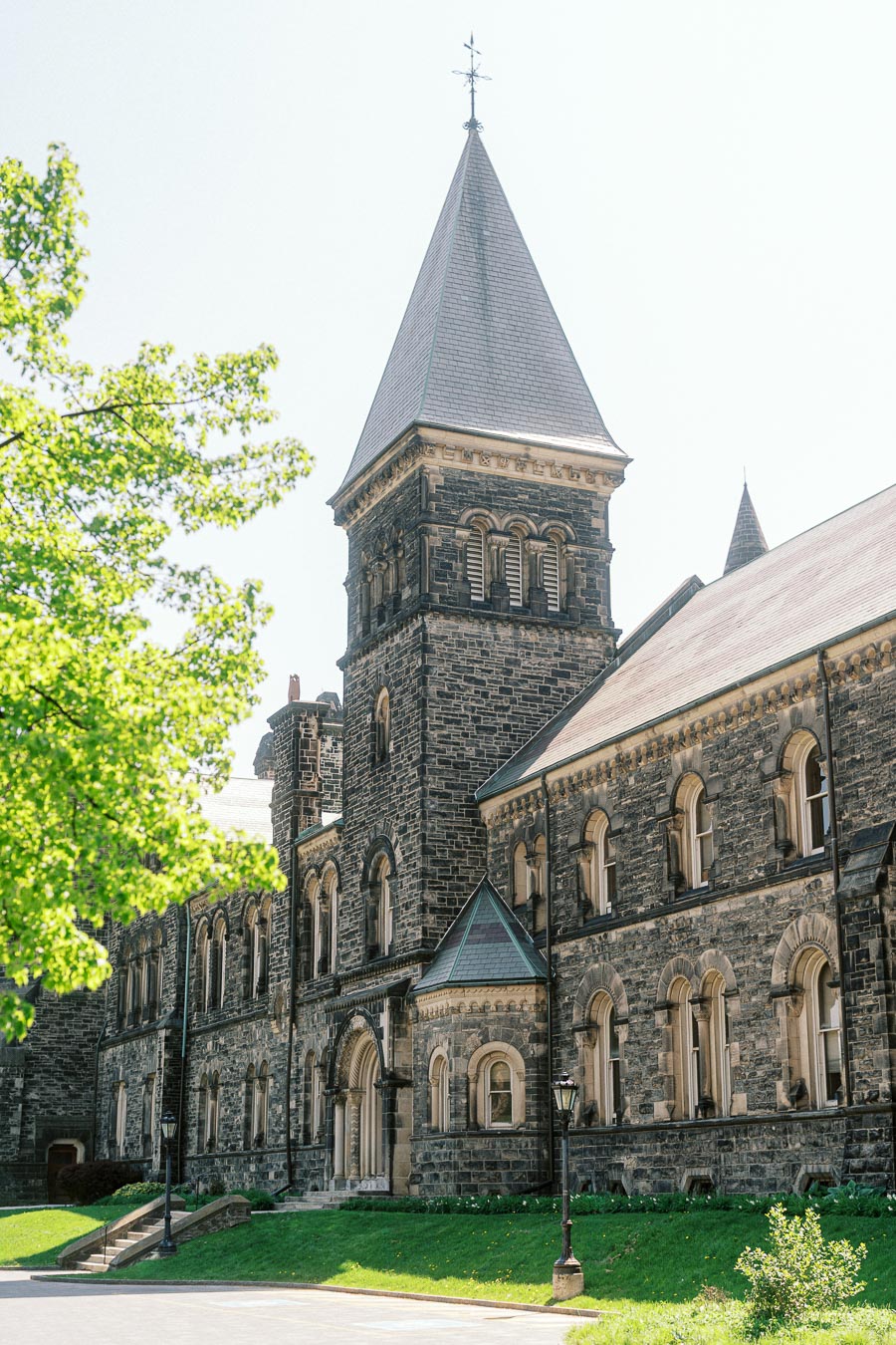 Historic stone building with tall spire and arched windows at University of Toronto, framed by spring greenery and bright sunlight.