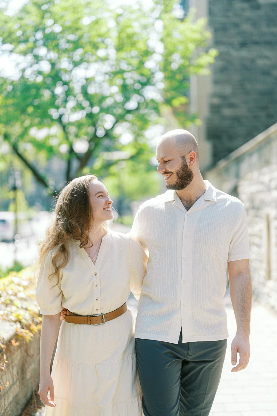 Engaged couple walking arm in arm, smiling at each other on a sunny spring day at the University of Toronto with greenery and stone walls in the background.