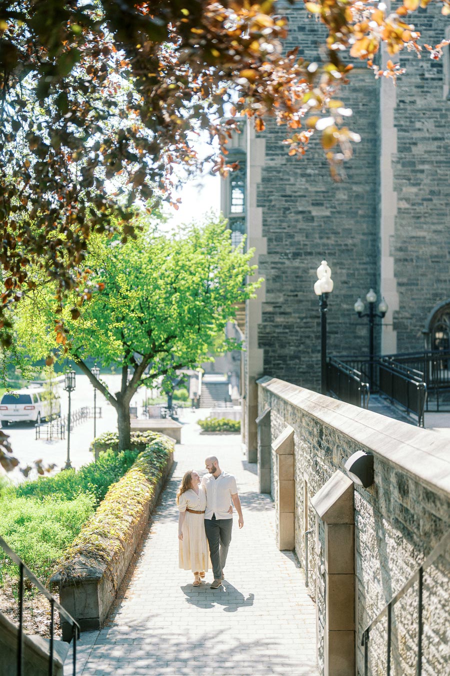 Couple walking together on a sunlit pathway at University of Toronto, framed by blooming trees and historic stone architecture in spring.