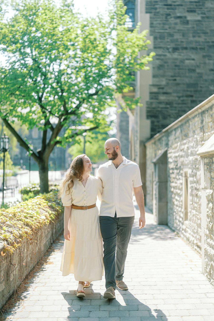 Couple walks arm in arm along a stone path at University of Toronto, surrounded by greenery and historic architecture in spring sunlight.