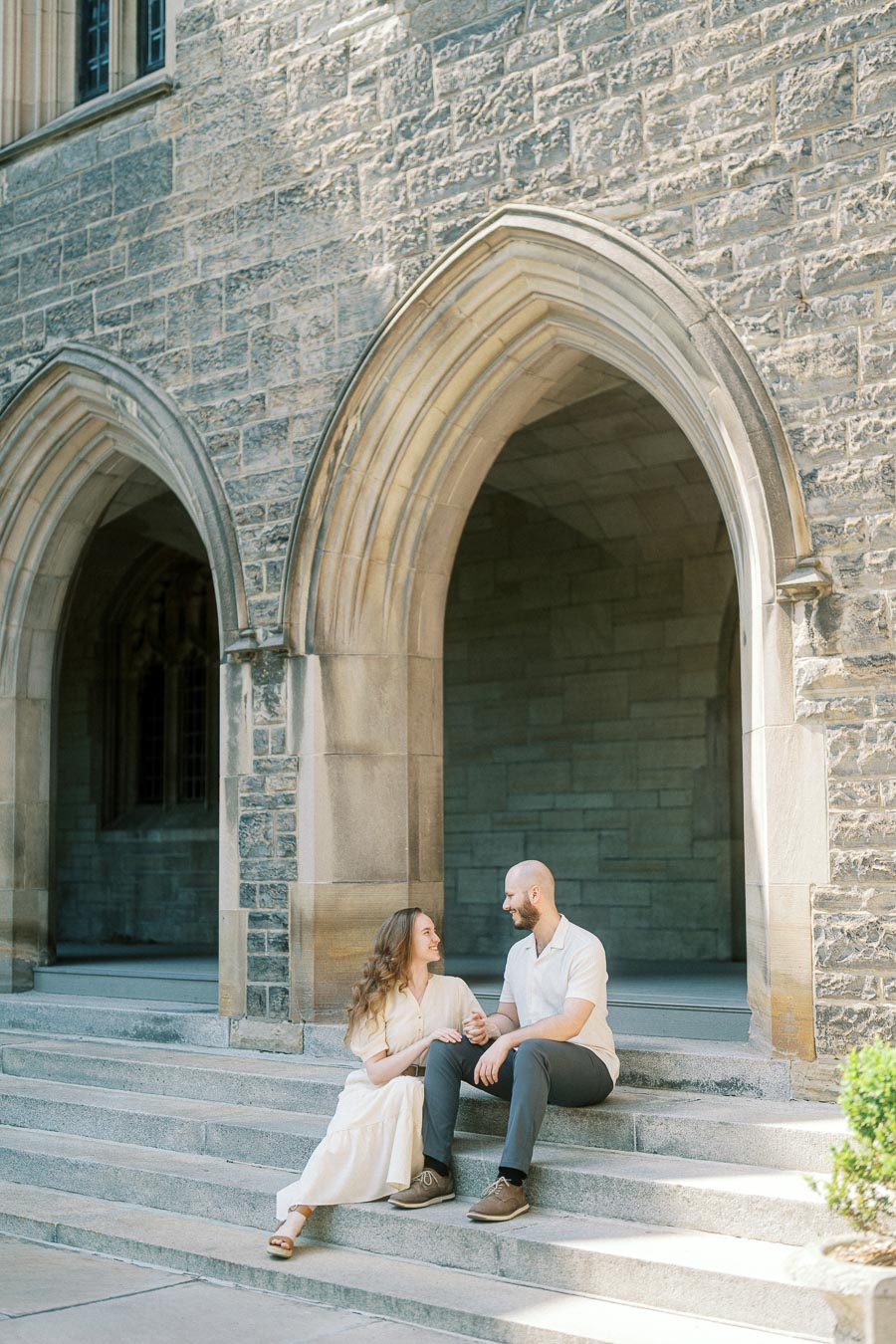 Couple sitting on stone steps under gothic arches at University of Toronto, holding hands and smiling in soft spring sunlight.