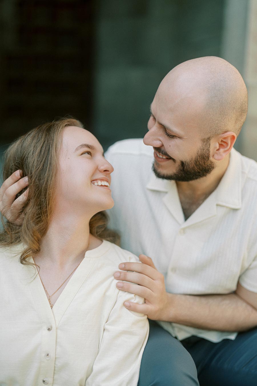 A couple smiling warmly at each other, sitting close together in soft natural light during a spring engagement session at the University of Toronto.