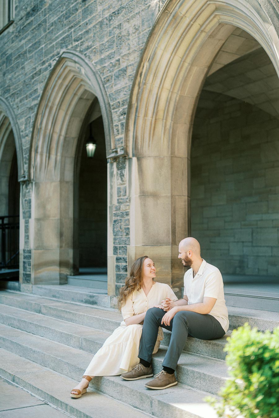 Engaged couple sits on stone steps beneath gothic arches at the University of Toronto, smiling at each other in soft spring sunlight.