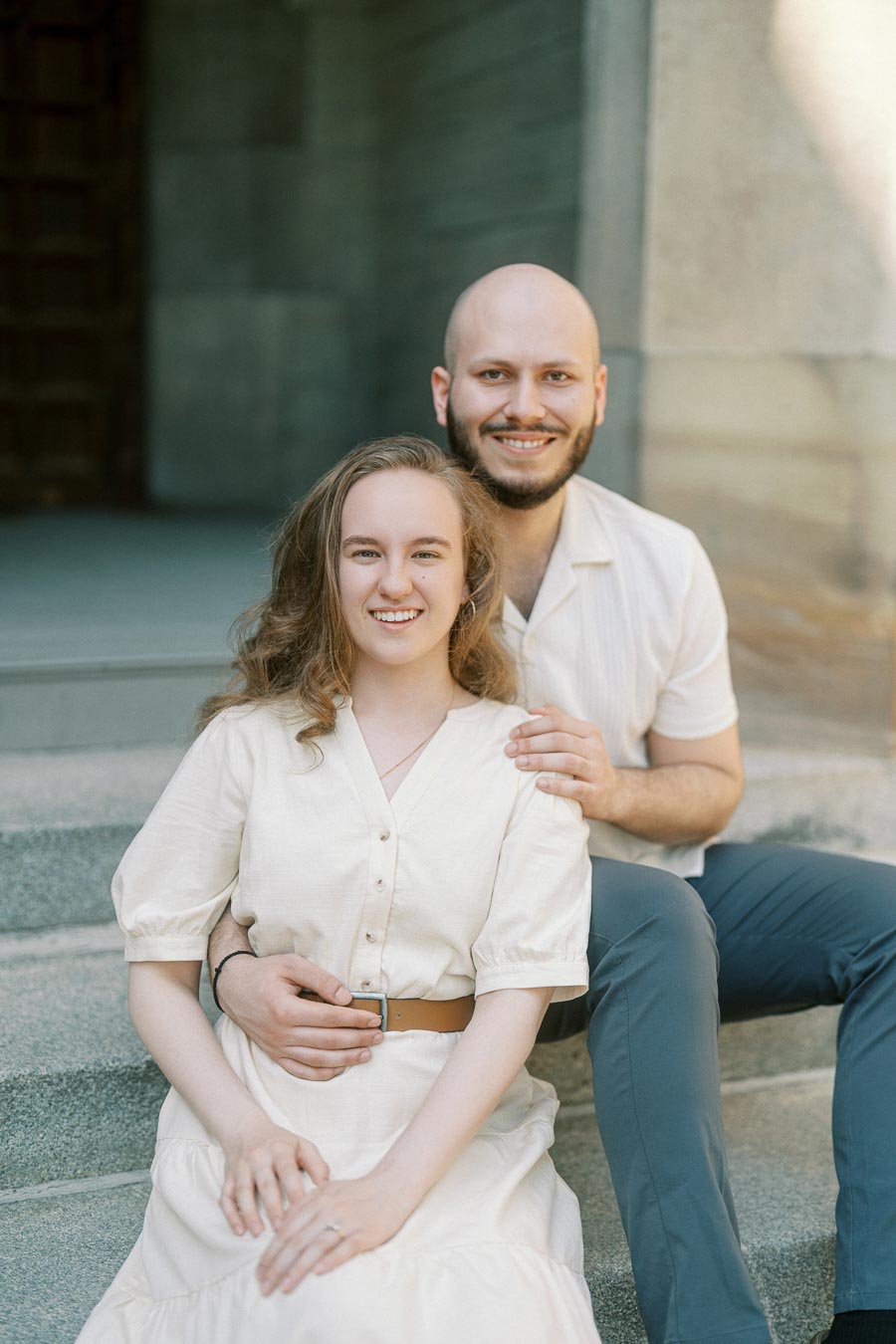 Couple sitting closely on stone steps, smiling warmly during a spring engagement session at the University of Toronto.