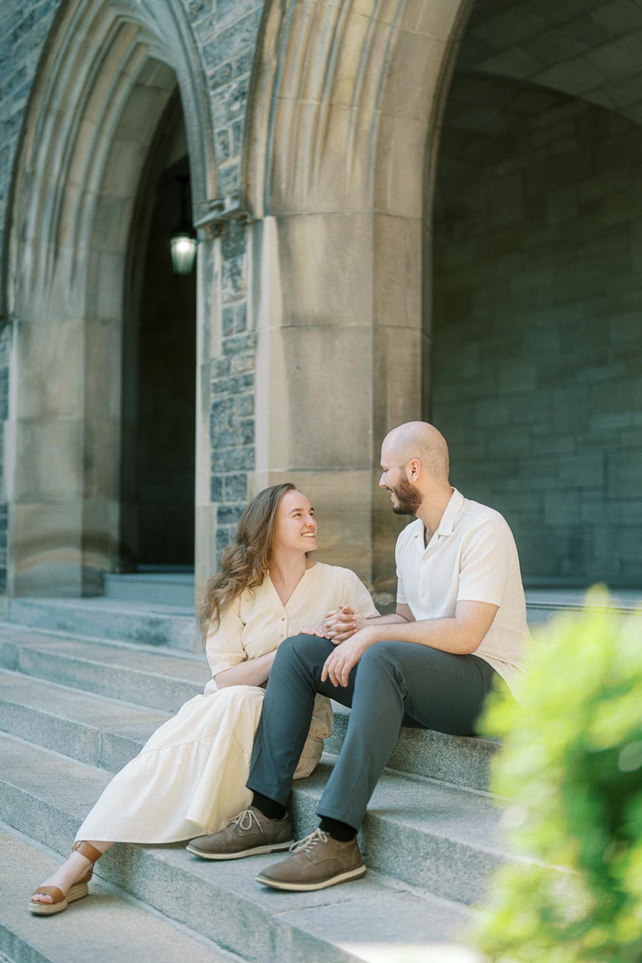 Couple sitting on stone steps beneath a University of Toronto archway, smiling at each other with spring greenery in the foreground.