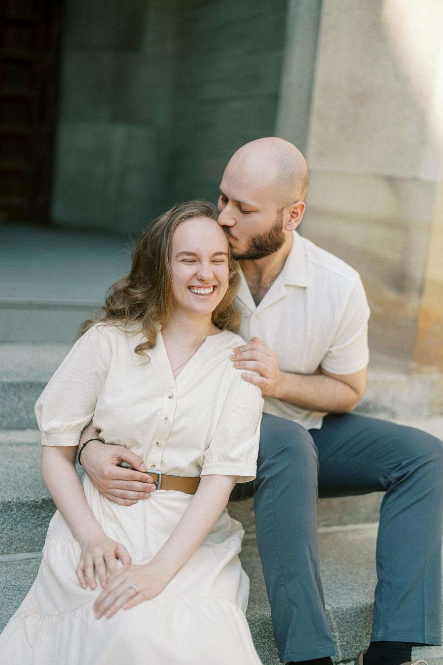 A couple sits on stone steps, the man gently kissing the woman's forehead as she smiles, surrounded by soft spring light at the University of Toronto.