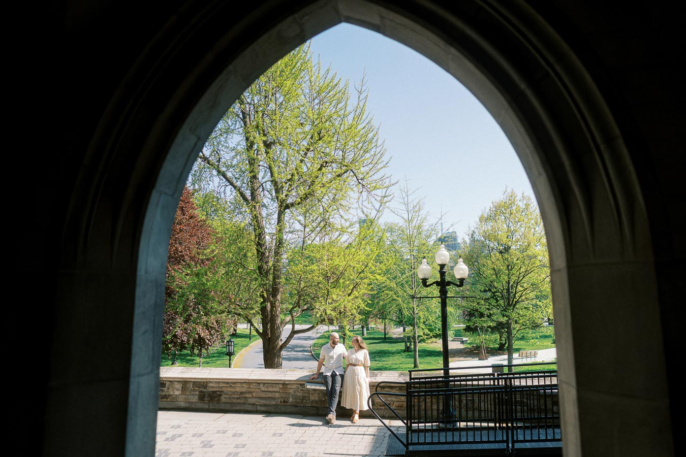 Couple standing beneath a stone archway at University of Toronto, framed by blooming spring trees and historic campus architecture.