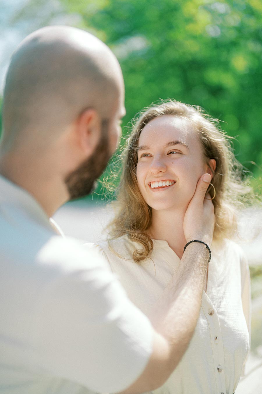 A smiling woman looks lovingly at her partner as he gently touches her face during a sunny spring engagement session at the University of Toronto.