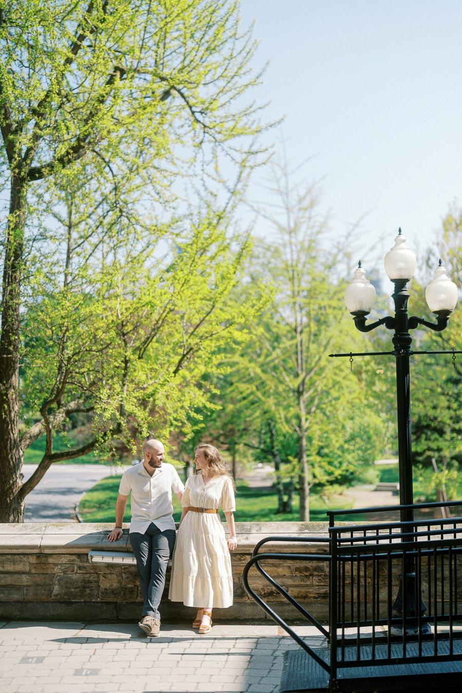Couple sitting together on a stone ledge at the University of Toronto, smiling at each other under blooming spring trees and historic lampposts.