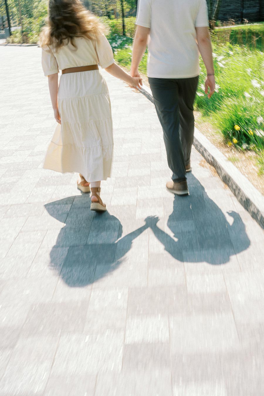 A couple walks hand in hand on a bright pathway at the University of Toronto, their shadows holding hands in the sunlight.