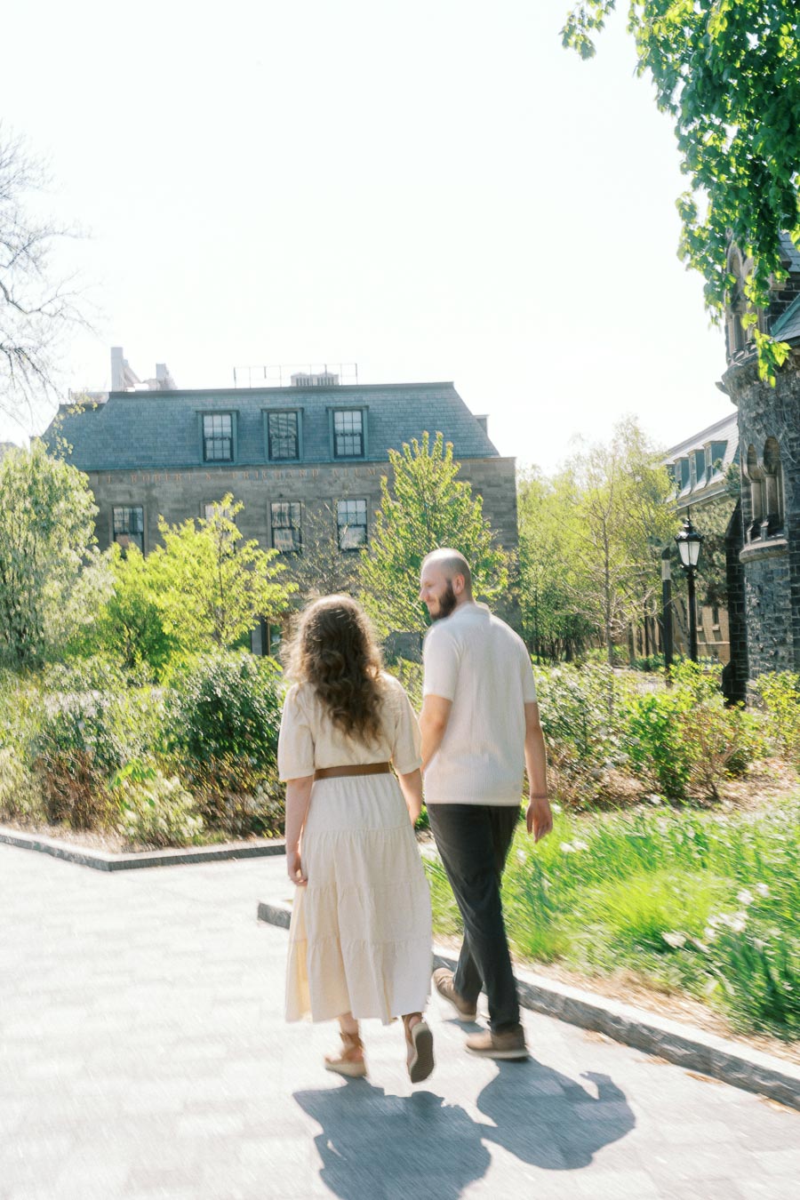 Romantic couple walks together on a sunlit path surrounded by greenery and historic campus buildings at the University of Toronto in spring.