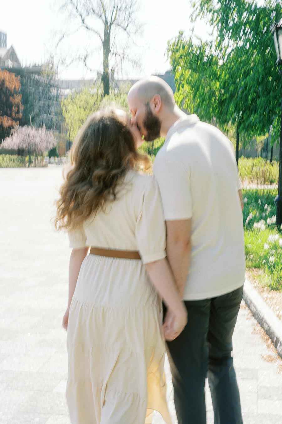 Couple kissing while walking hand in hand on a sunny path at the University of Toronto, surrounded by greenery and spring blooms.