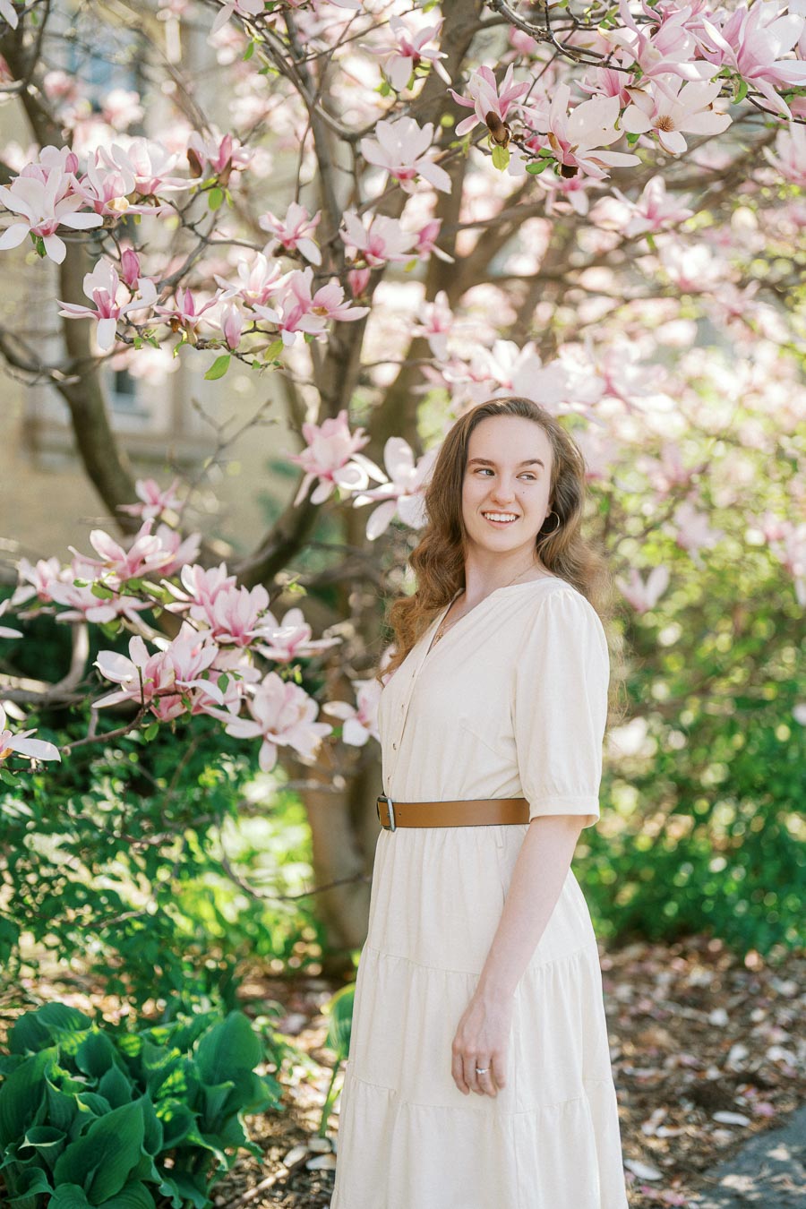 Woman in a cream dress smiles beneath blooming pink magnolia branches on a sunny spring day at University of Toronto.