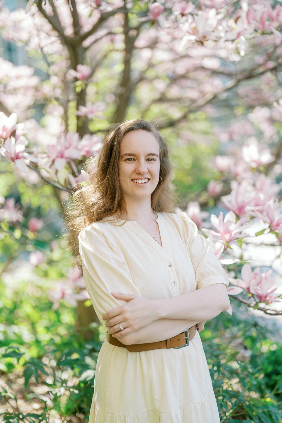 Woman in a cream dress smiles with arms crossed, surrounded by blooming pink magnolia flowers in spring sunlight at University of Toronto.