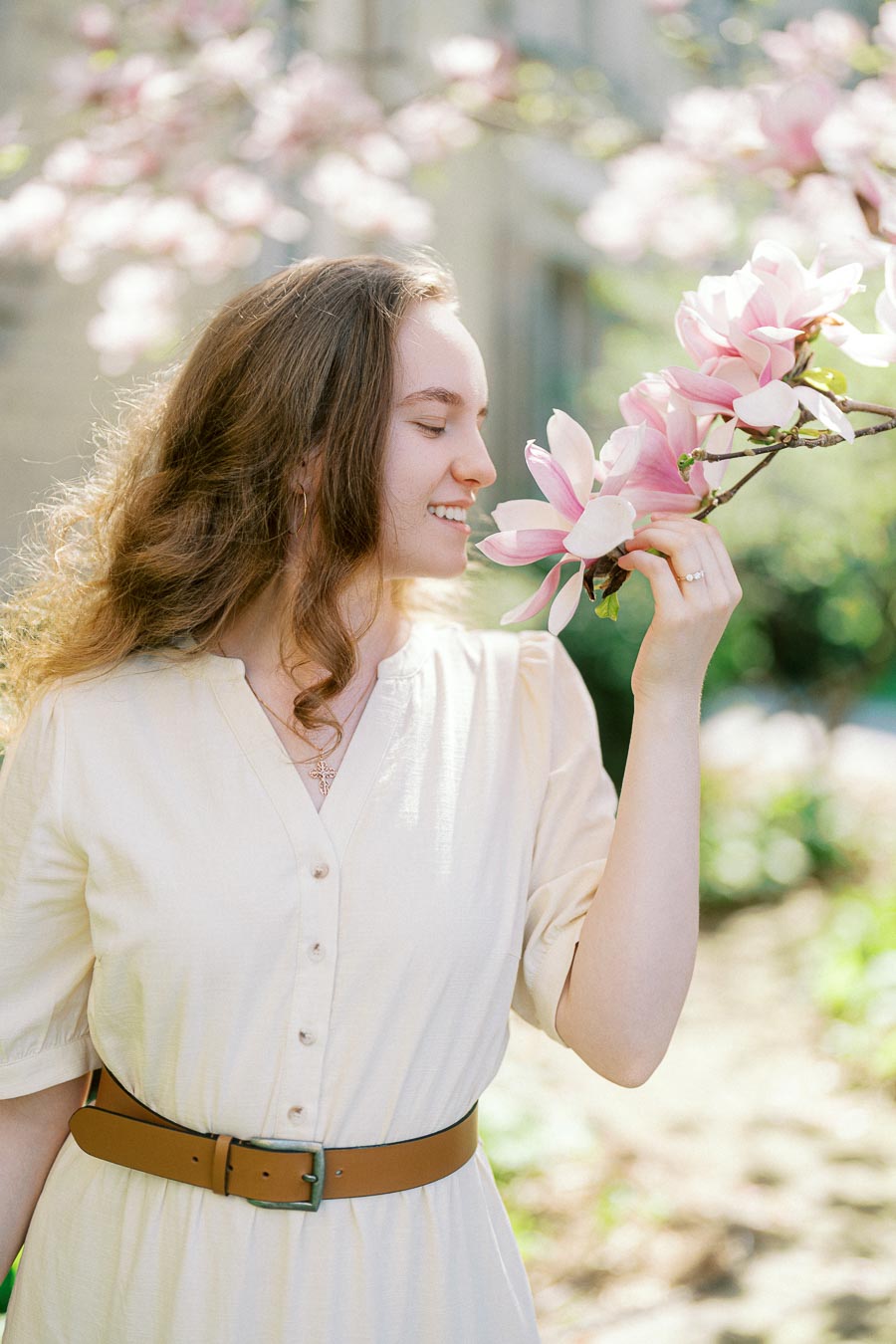 Bride-to-be in a cream dress smiles while admiring pink magnolia blossoms on a sunny spring day at University of Toronto.