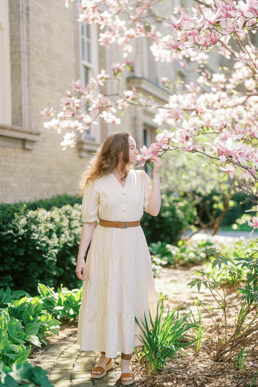 Woman in a cream dress smelling pink magnolia blossoms in a sunlit garden at the University of Toronto campus in spring.