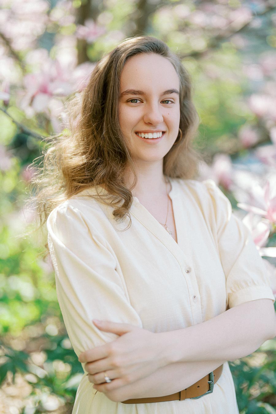 Woman with wavy hair in a cream dress smiles in front of blooming pink flowers and greenery at the University of Toronto in spring.