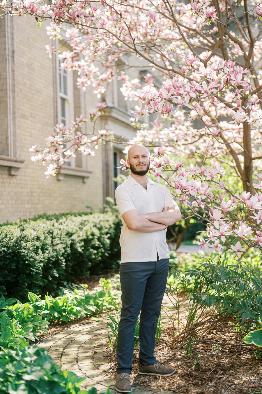 Man with arms crossed stands under a blooming magnolia tree in spring at University of Toronto, with historic architecture in the background.