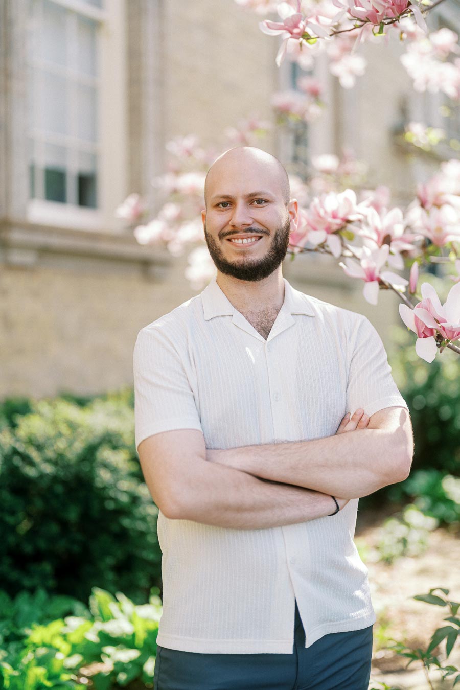 Smiling man with a beard stands with arms crossed under blooming pink magnolia trees at University of Toronto in spring sunlight.