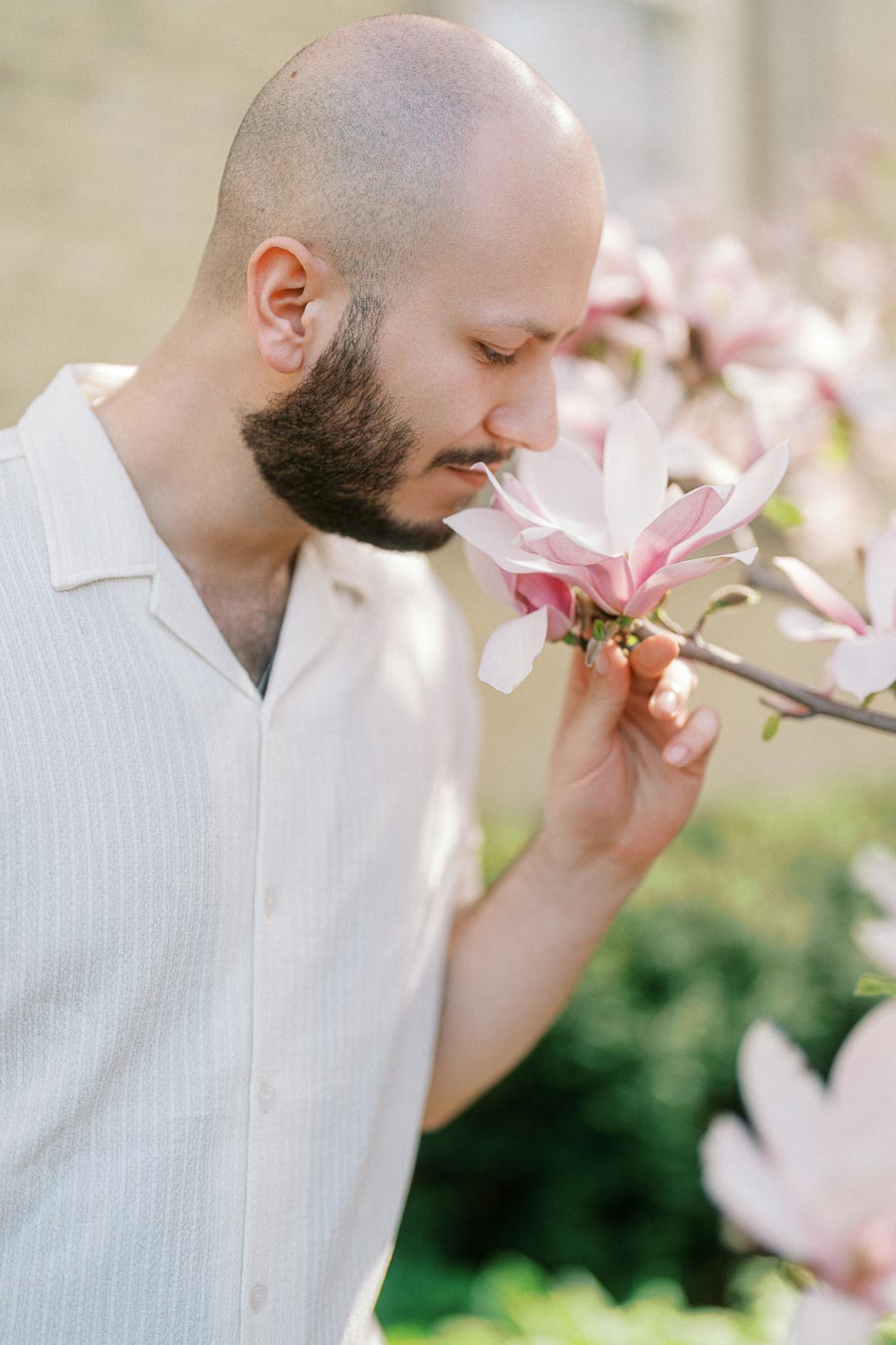 Man in a light shirt smelling a pink and white magnolia blossom on campus during a spring engagement shoot in Toronto.