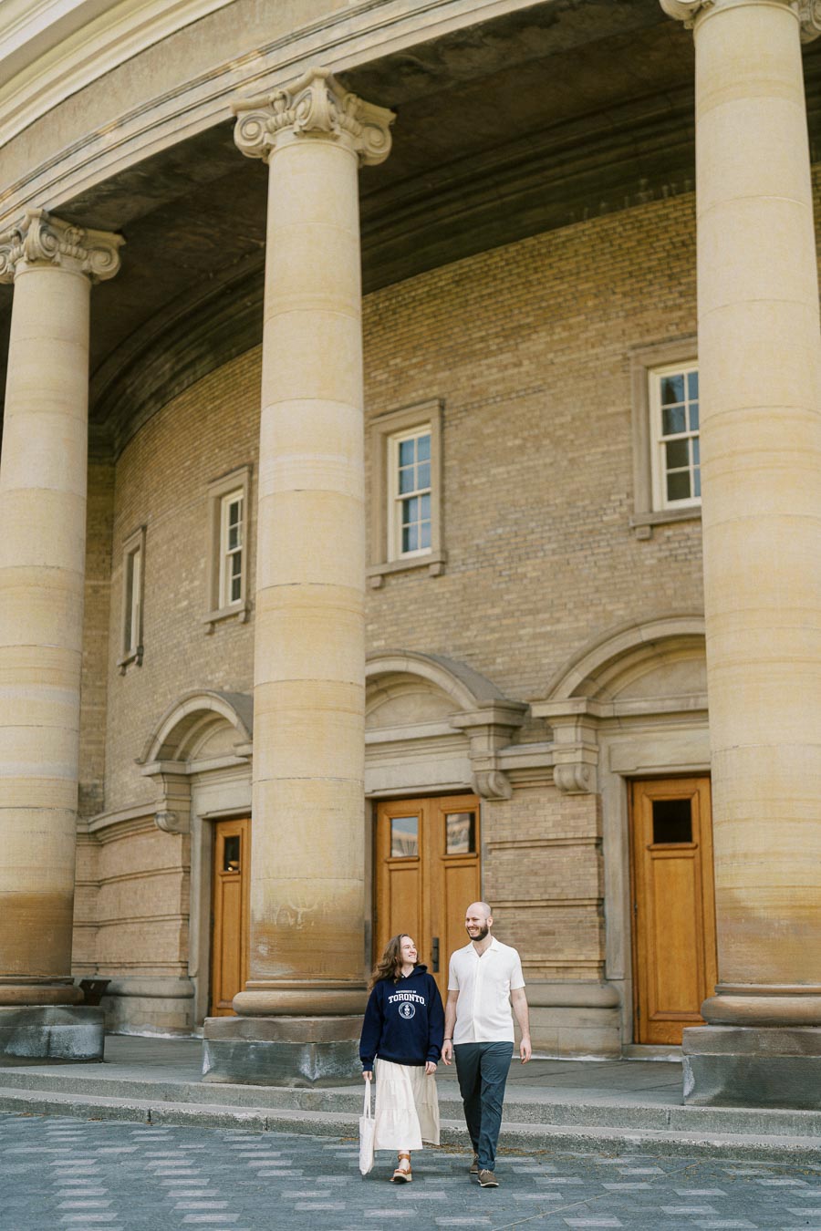 A couple walks hand in hand beneath historic stone columns at the University of Toronto, smiling and enjoying a sunny spring day.
