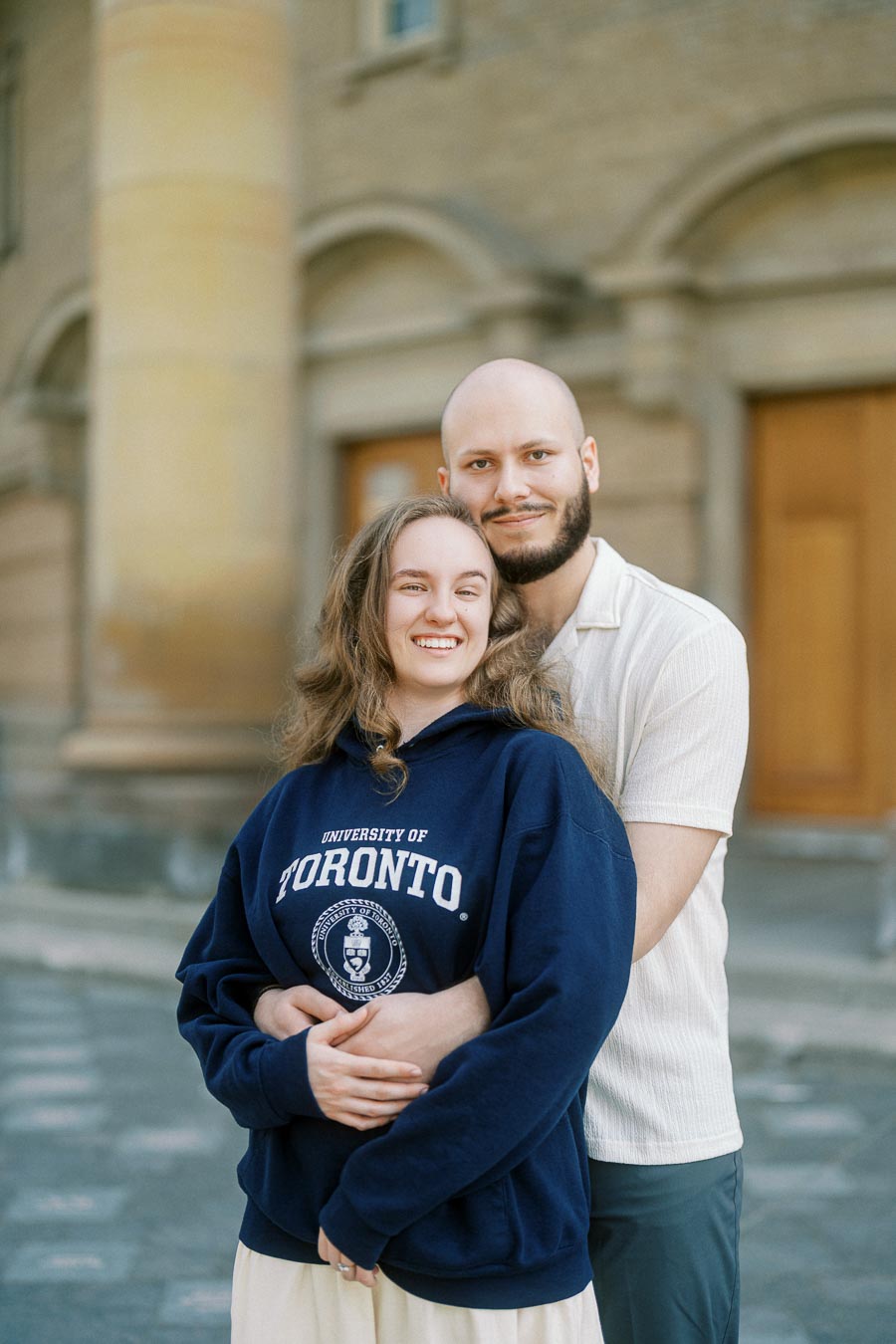 Smiling couple embraces in front of University of Toronto building, capturing a romantic spring engagement moment on campus.