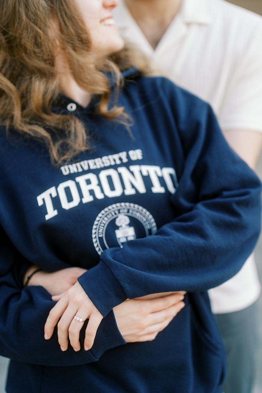 Close-up of a smiling engaged couple embracing, with the woman wearing a University of Toronto sweatshirt and showing her engagement ring.