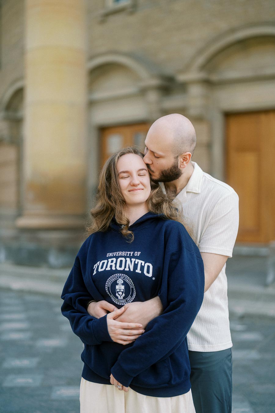 Couple embraces as one partner kisses the other's forehead, standing in front of historic University of Toronto columns in spring.