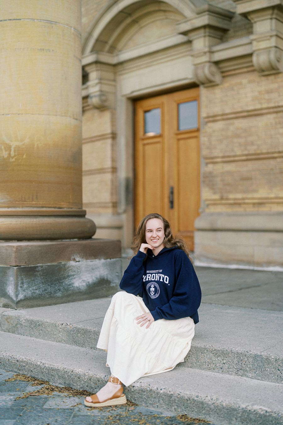 Woman wearing a University of Toronto hoodie sits on stone steps in front of historic campus architecture, smiling on a sunny spring day.