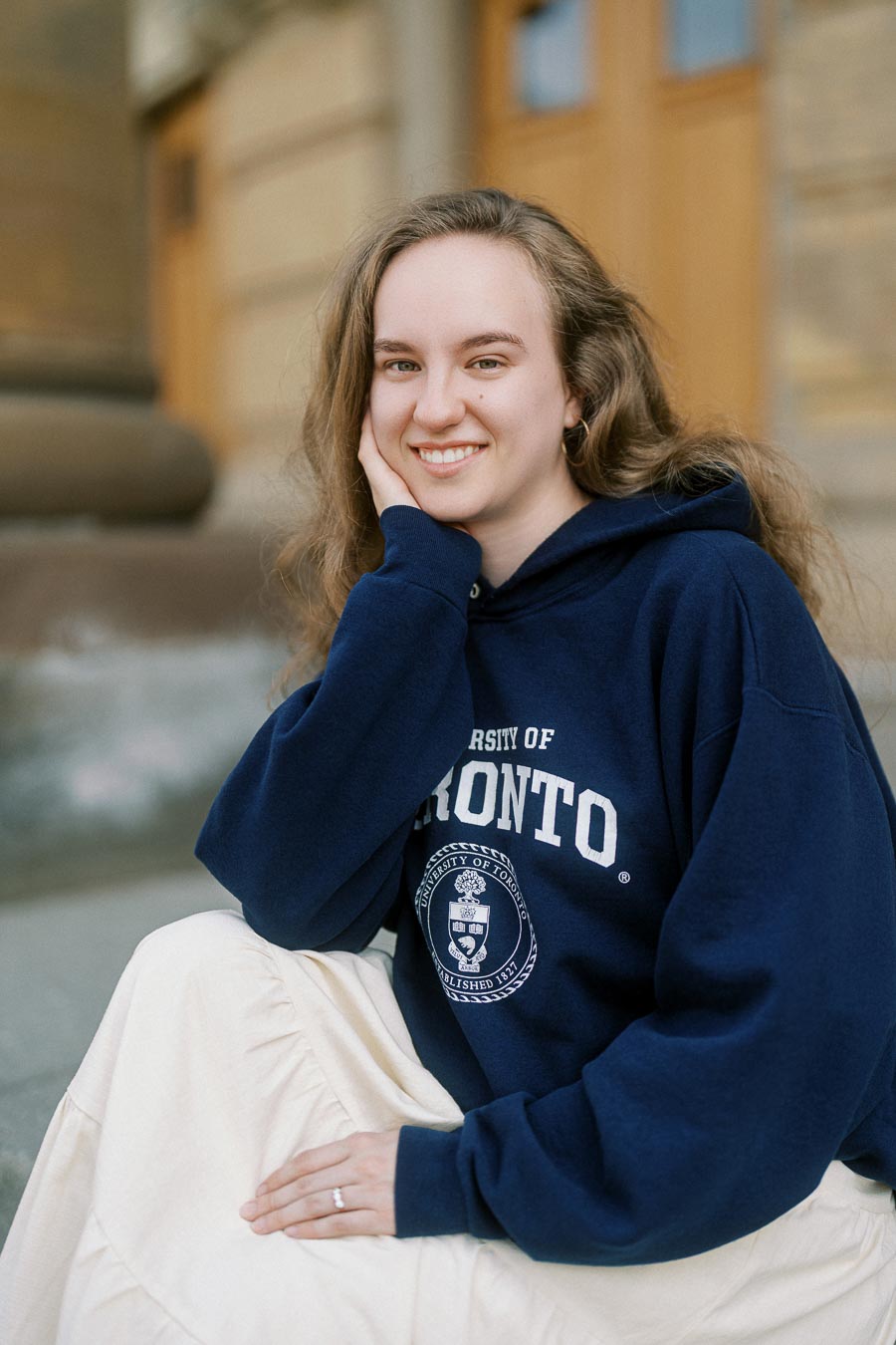 Maryanna smiles while seated on the University of Toronto steps, wearing a navy UofT hoodie and displaying her engagement ring.