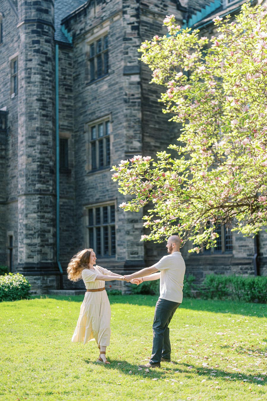 Couple laughing and holding hands while dancing on a sunny lawn beneath blooming trees at the University of Toronto in spring.