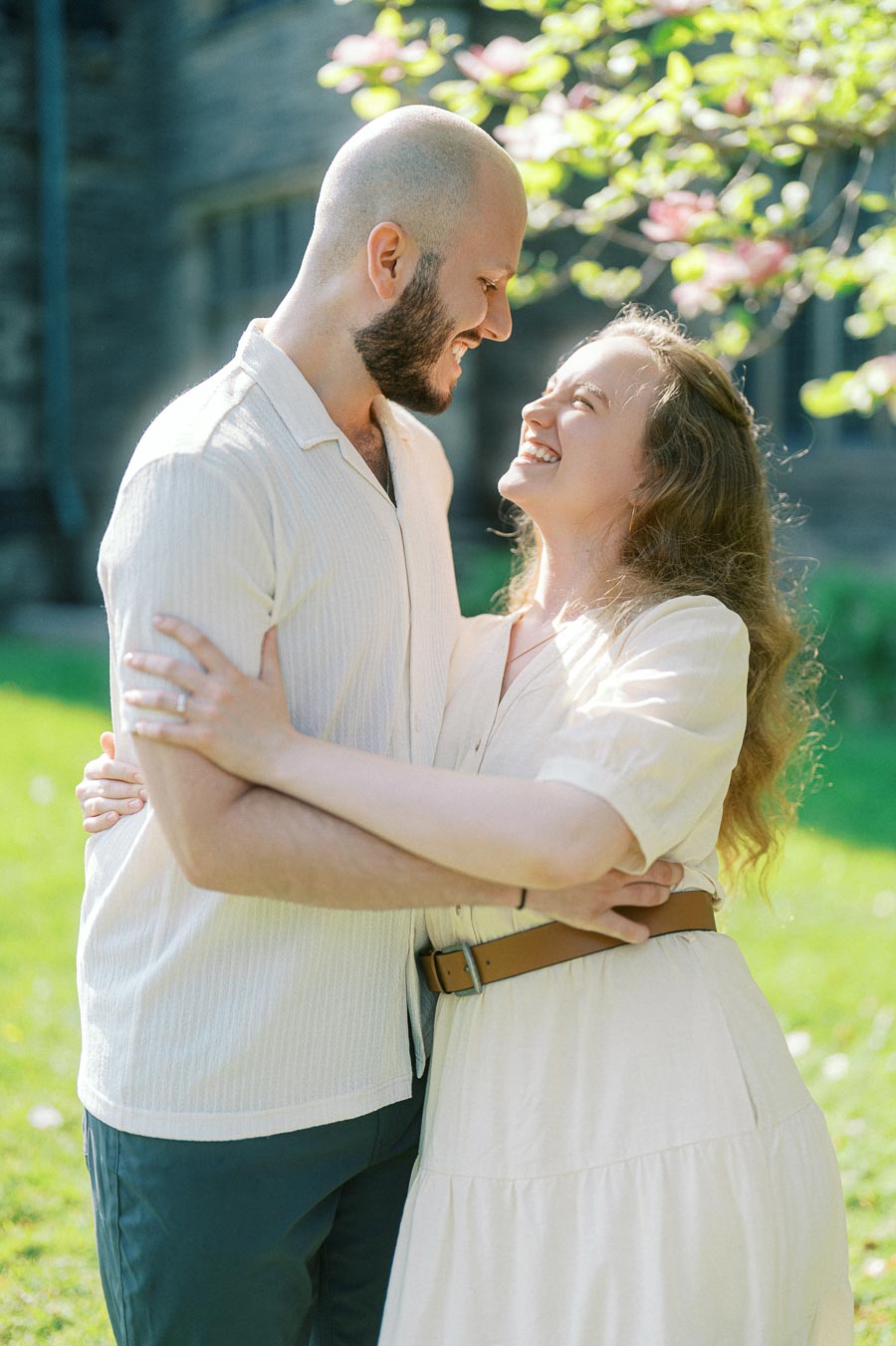 Engaged couple smiling and embracing under blooming spring branches on the University of Toronto campus, sunlight highlighting their happiness.