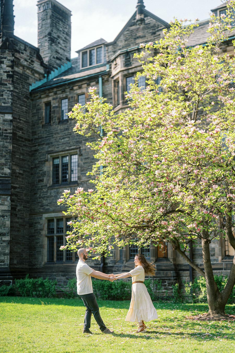 Engaged couple holding hands and spinning beneath a blooming tree on the University of Toronto campus in spring sunlight.