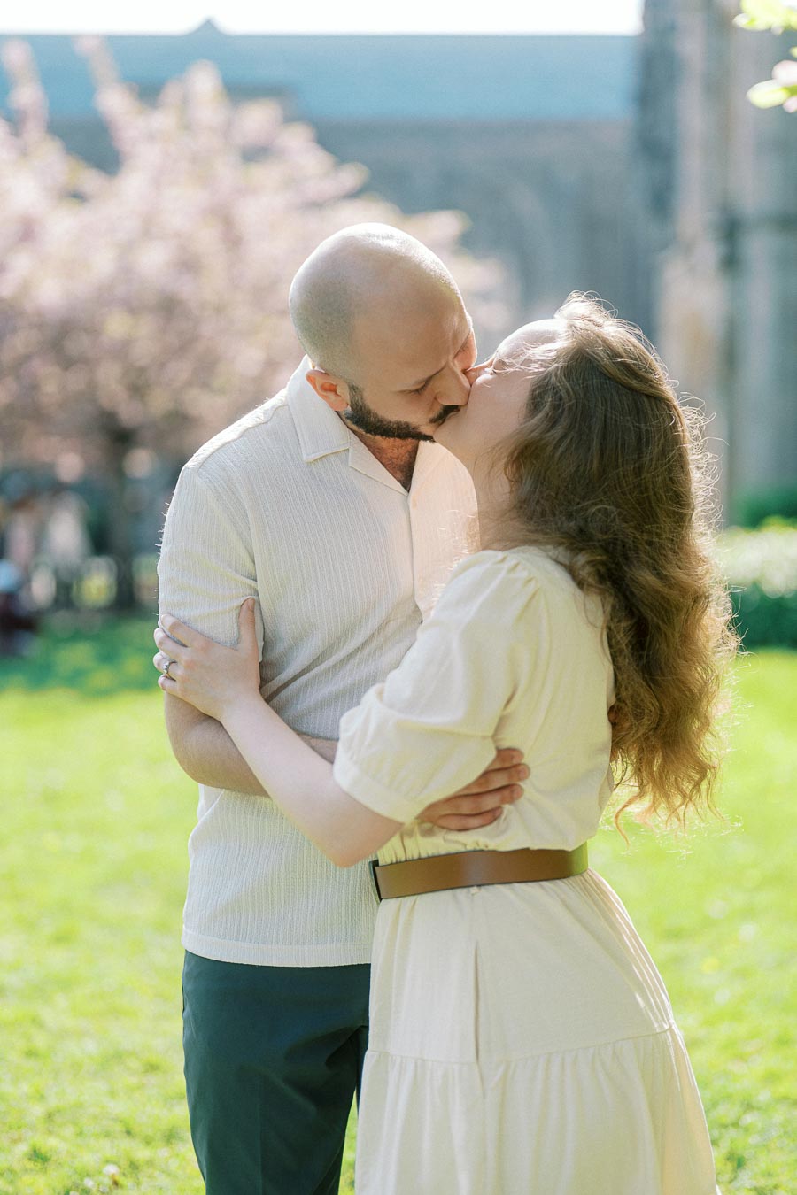 Couple shares a tender kiss in a sunlit University of Toronto courtyard with blooming spring trees and historic campus buildings in the background.