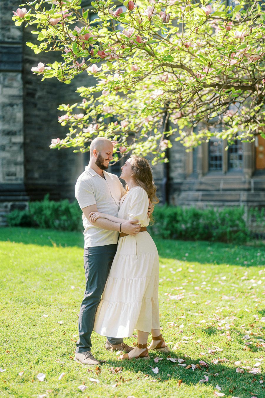 Couple embracing and smiling under a blooming magnolia tree on the University of Toronto campus in spring, with sunlight and historic stone building in the background.