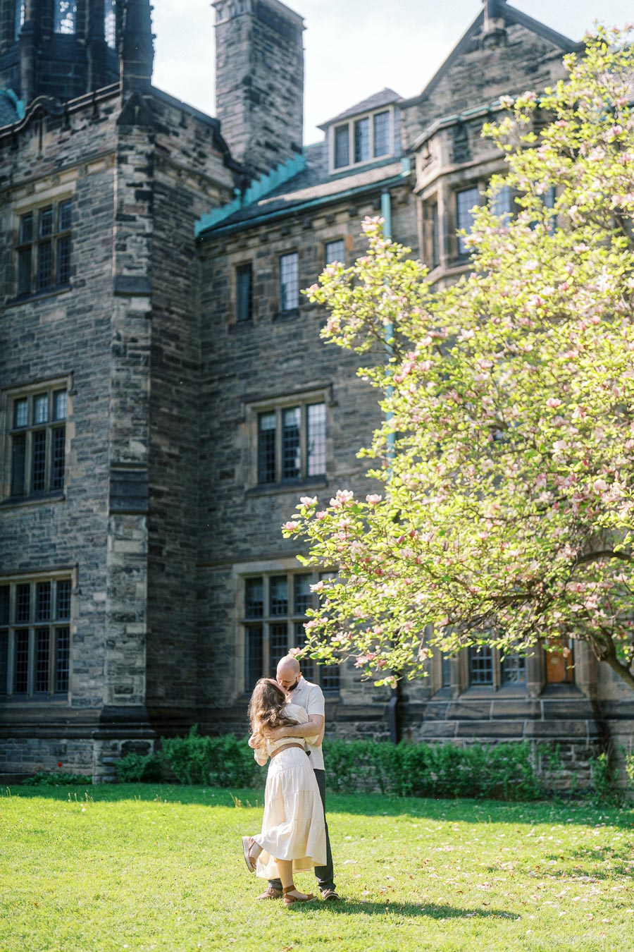 Couple embracing on a sunny lawn in front of historic stone building and blooming tree at University of Toronto in spring.