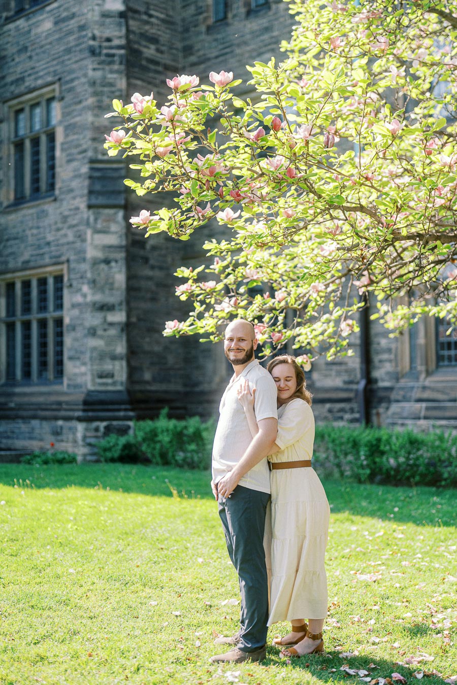 A couple stands embracing under a blooming tree on a sunny spring day, with historic stone architecture in the background at the University of Toronto.