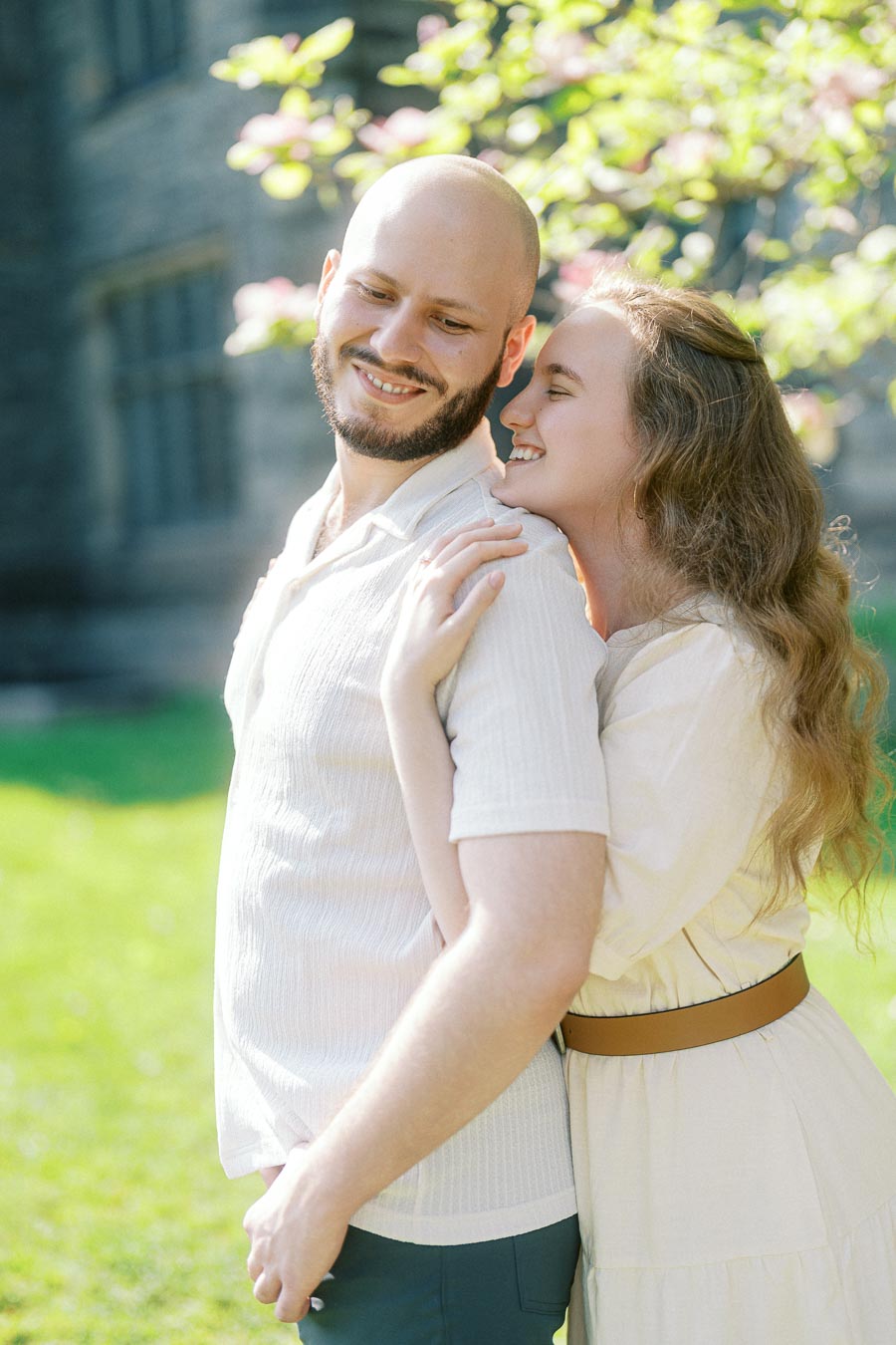 Couple smiling and embracing outdoors in spring sunlight with blooming trees at the University of Toronto campus.