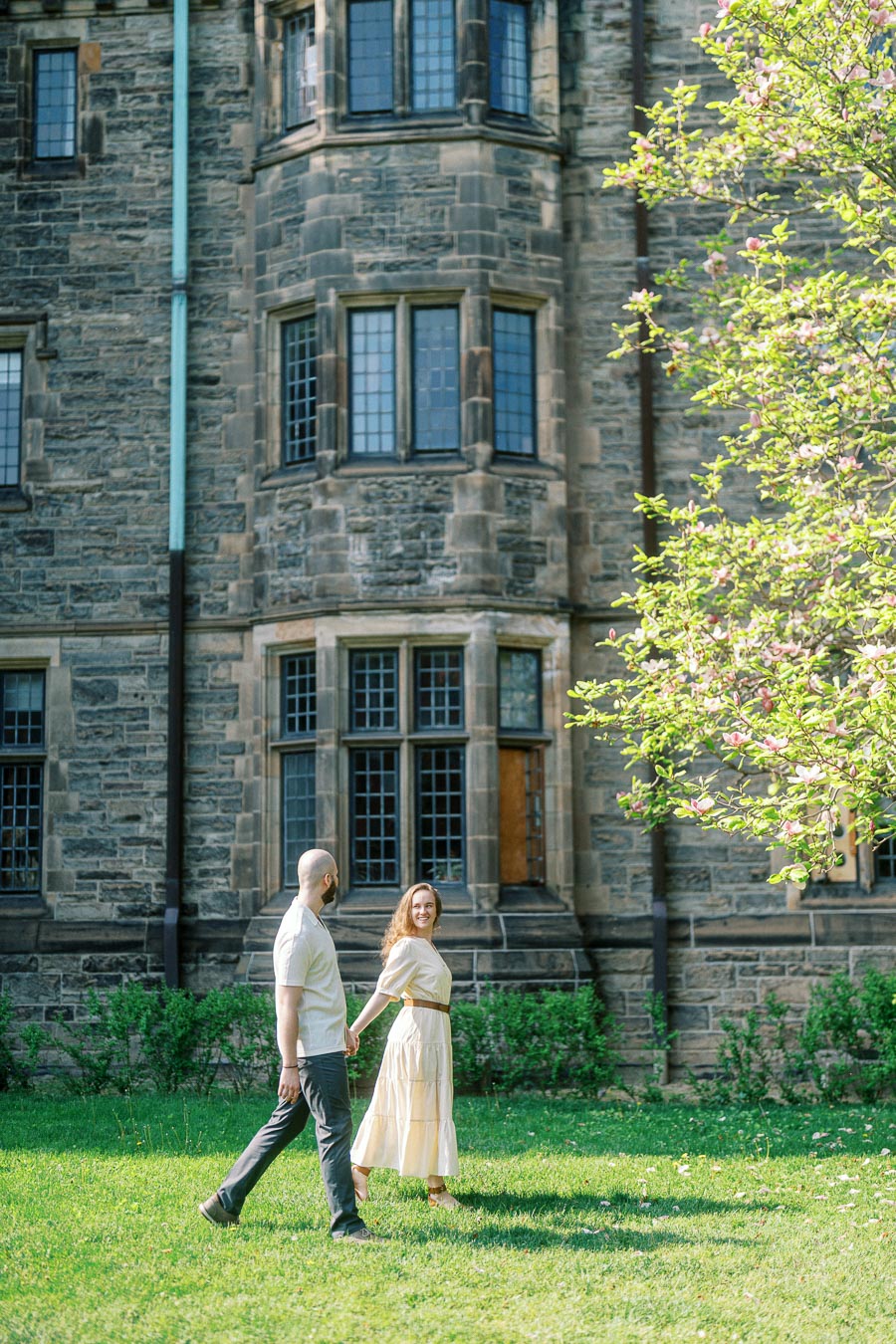 Couple holding hands and smiling while walking on a sunny lawn in front of historic stone building at University of Toronto in spring.