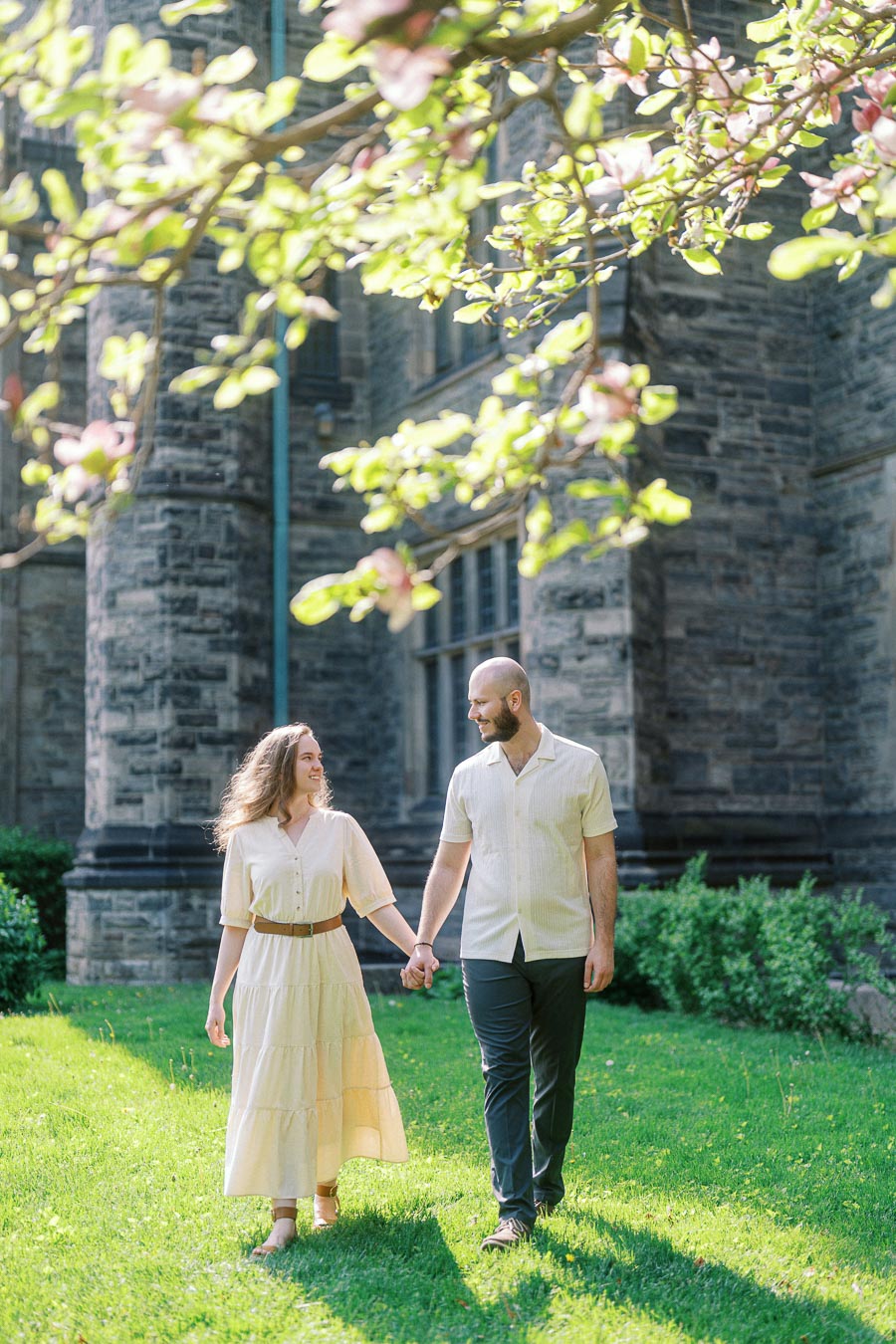 Couple walking hand-in-hand on green lawn beneath blooming tree branches at University of Toronto, with historic stone building in the background.