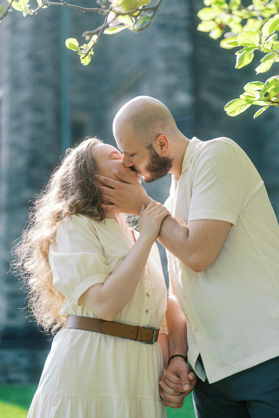 A couple shares a tender kiss while holding hands in spring sunlight at the University of Toronto, with lush greenery and stone architecture in the background.