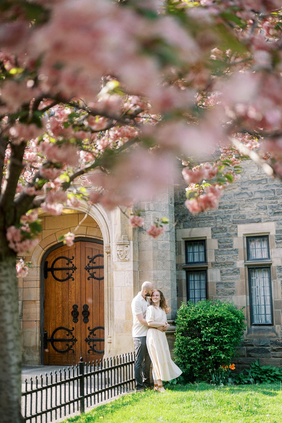 Couple embraces under pink cherry blossoms in front of a historic stone building at University of Toronto during a sunny spring engagement session.