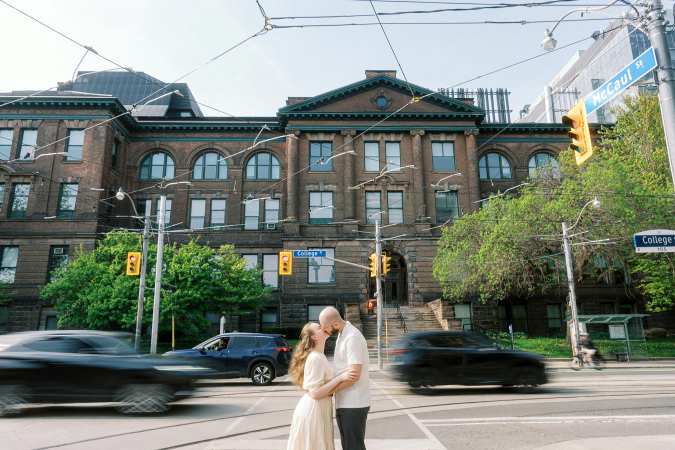 Couple embraces and kisses at a city intersection in front of the University of Toronto, with cars passing and spring greenery around them.
