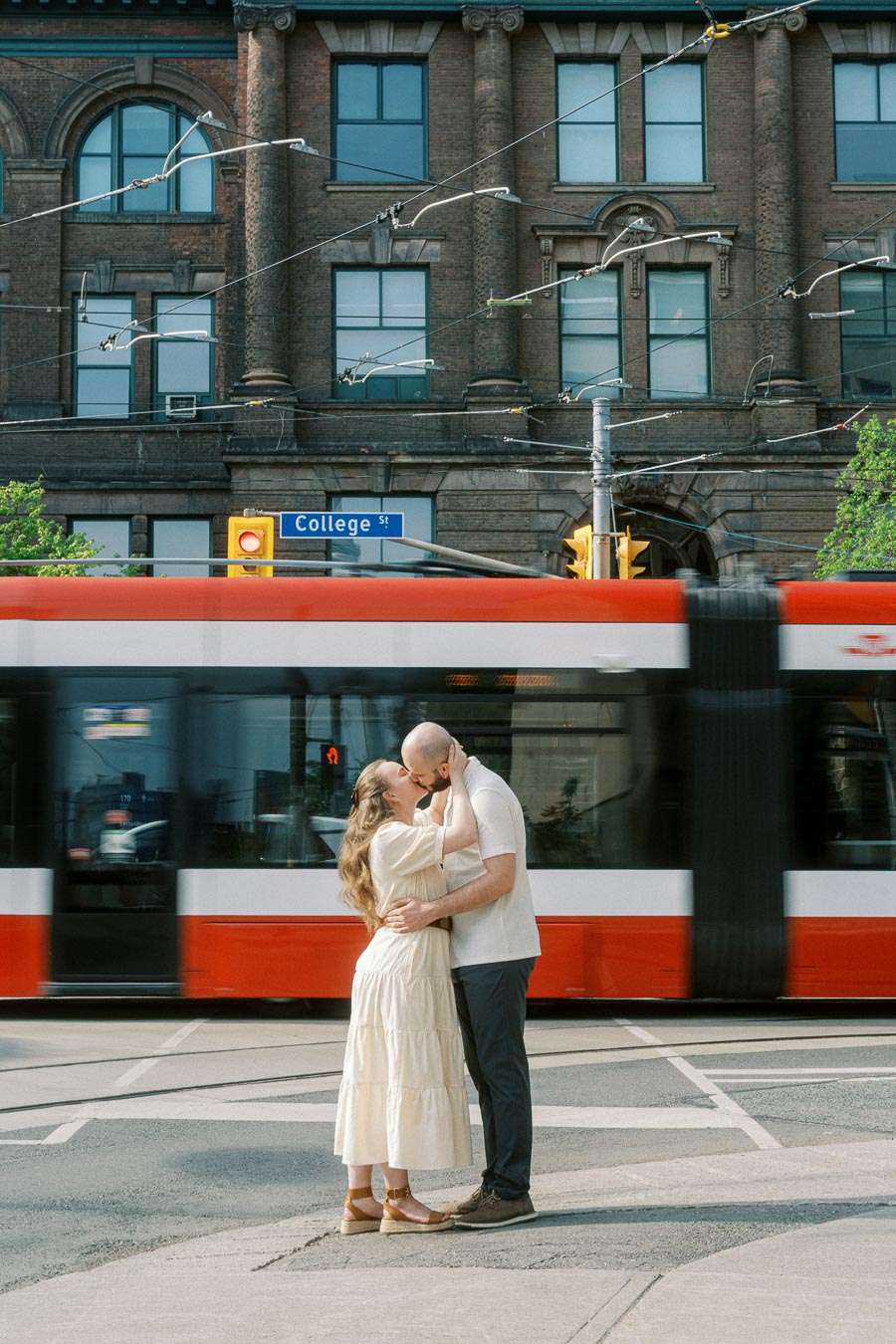 A couple embraces and kisses on a Toronto street corner as a red and white streetcar passes, with historic University of Toronto buildings in the background.