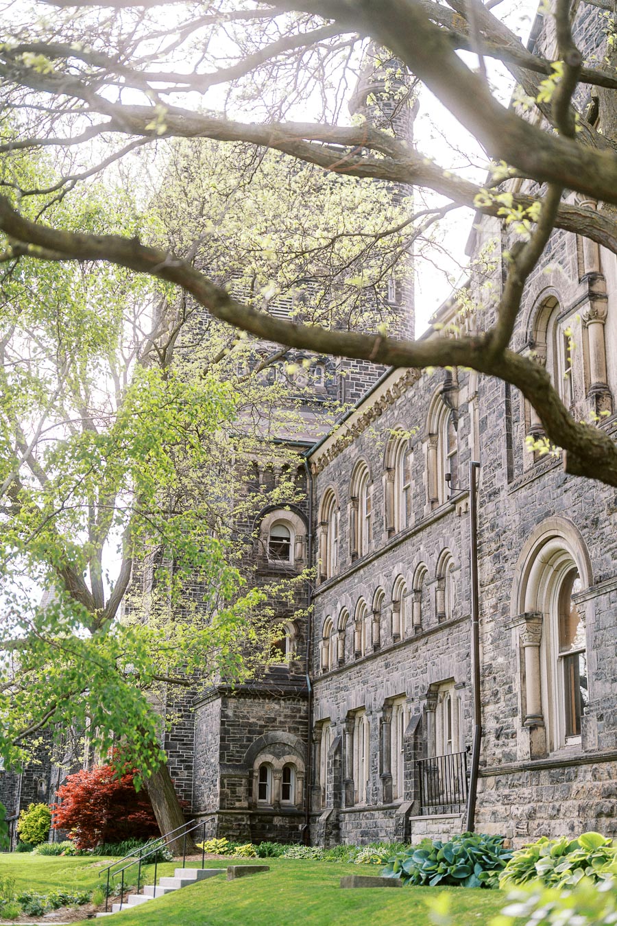 Stone university building with arched windows surrounded by green trees and landscaped grass at University of Toronto St. George campus.