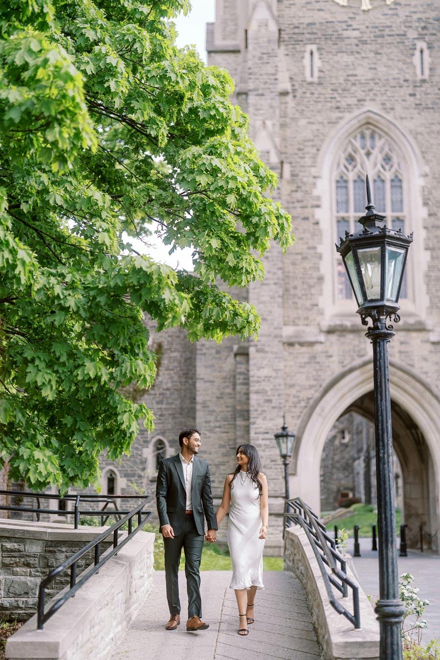 Tej and Priyana walk hand in hand along a stone pathway beneath green trees and a historic archway at the University of Toronto.