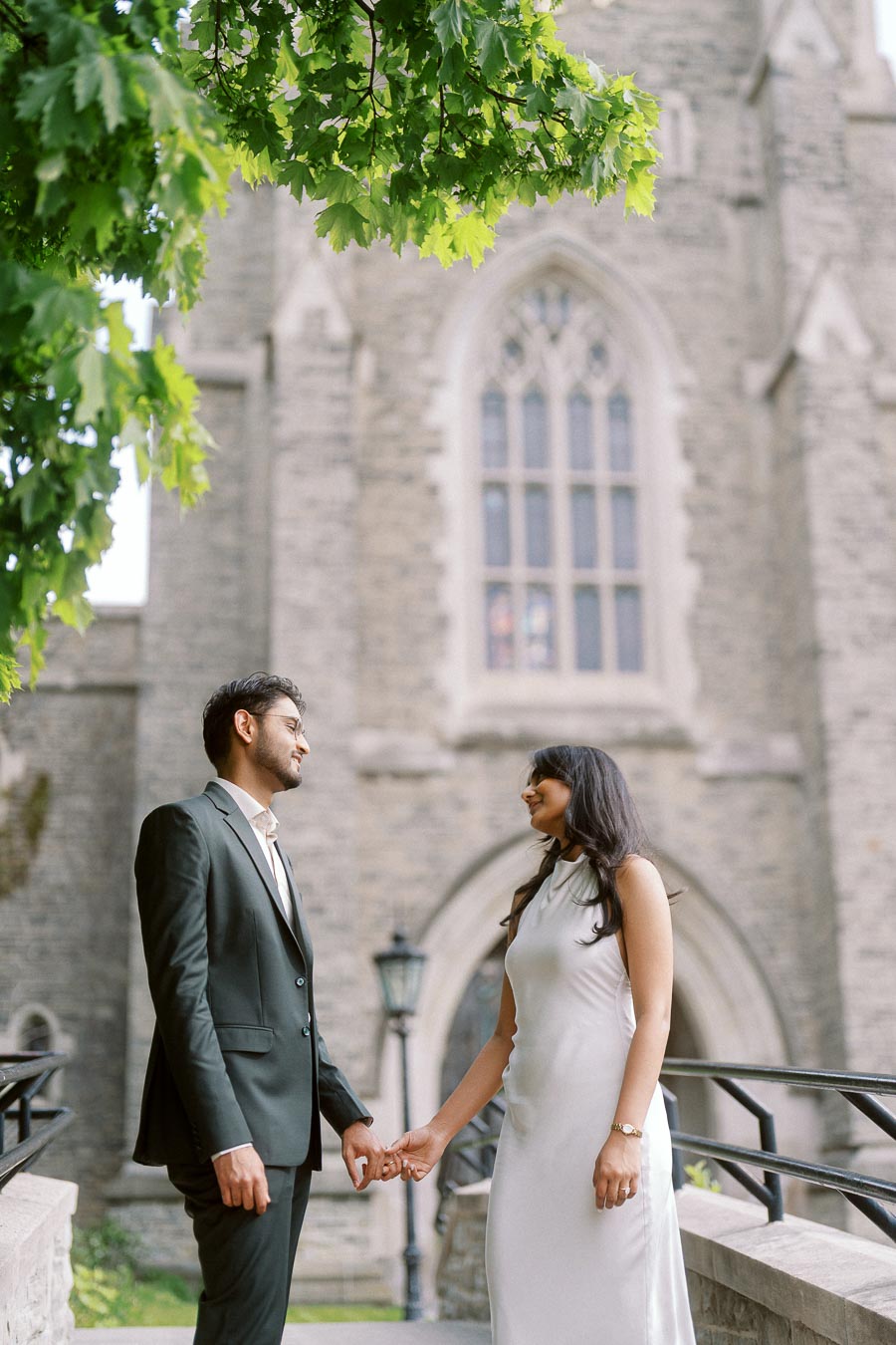 A couple stands holding hands beneath leafy branches in front of a historic stone building at University of Toronto.