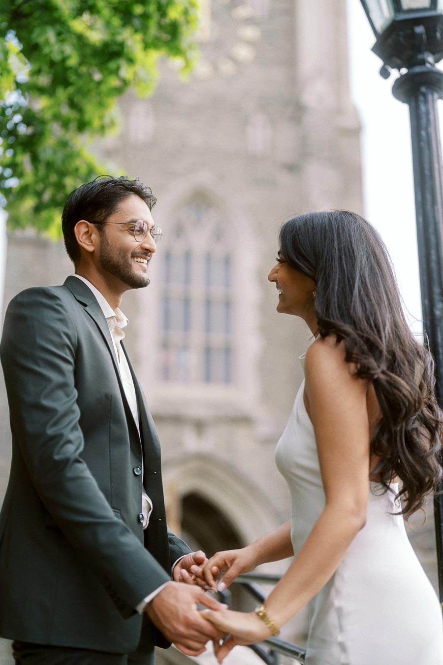 Tej and Priyana stand facing each other, holding hands and smiling, with a stone building at the University of Toronto behind them.