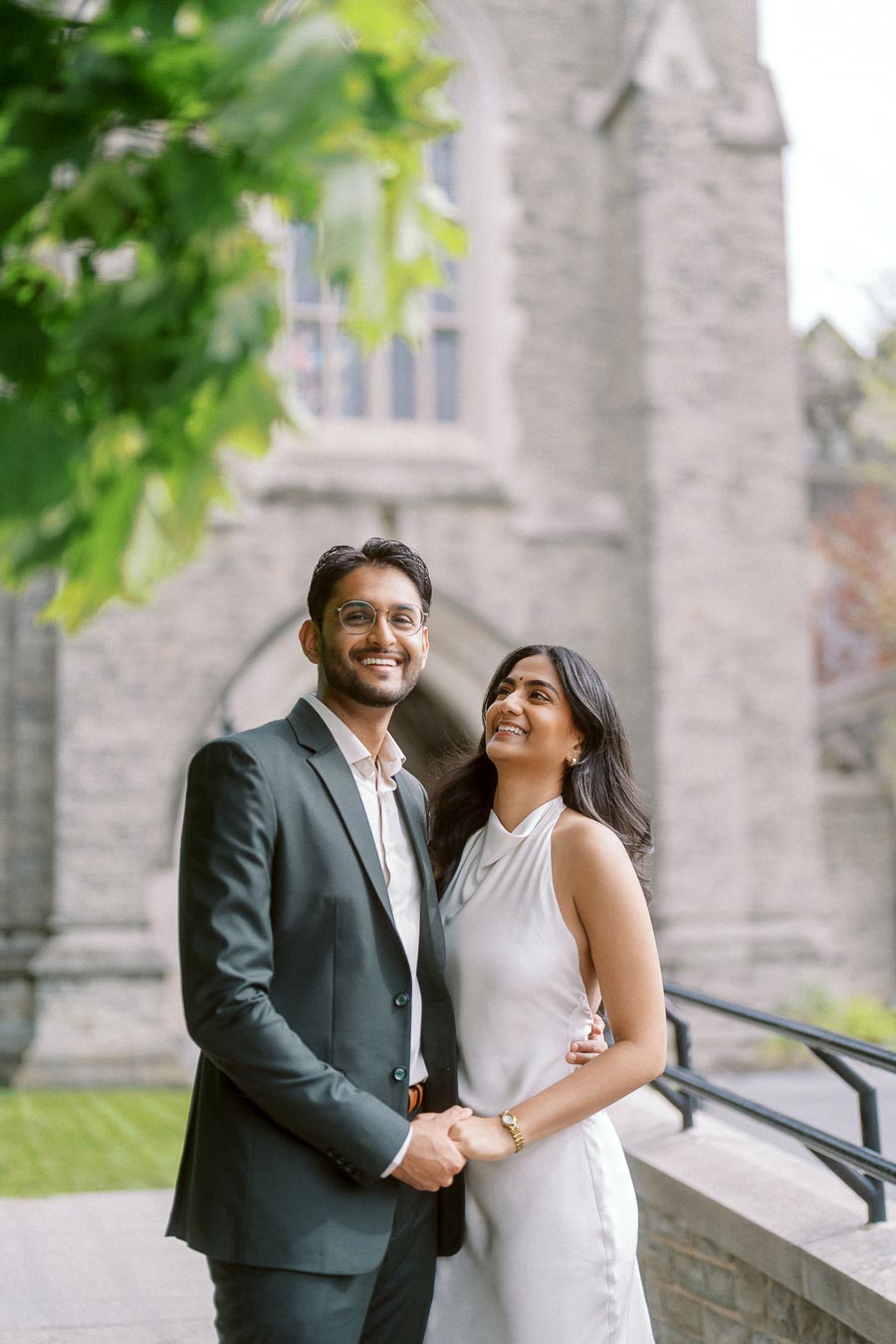 A couple, Tej and Priyana, stand closely together and smile in front of a stone building at the University of Toronto, with green leaves in the foreground.
