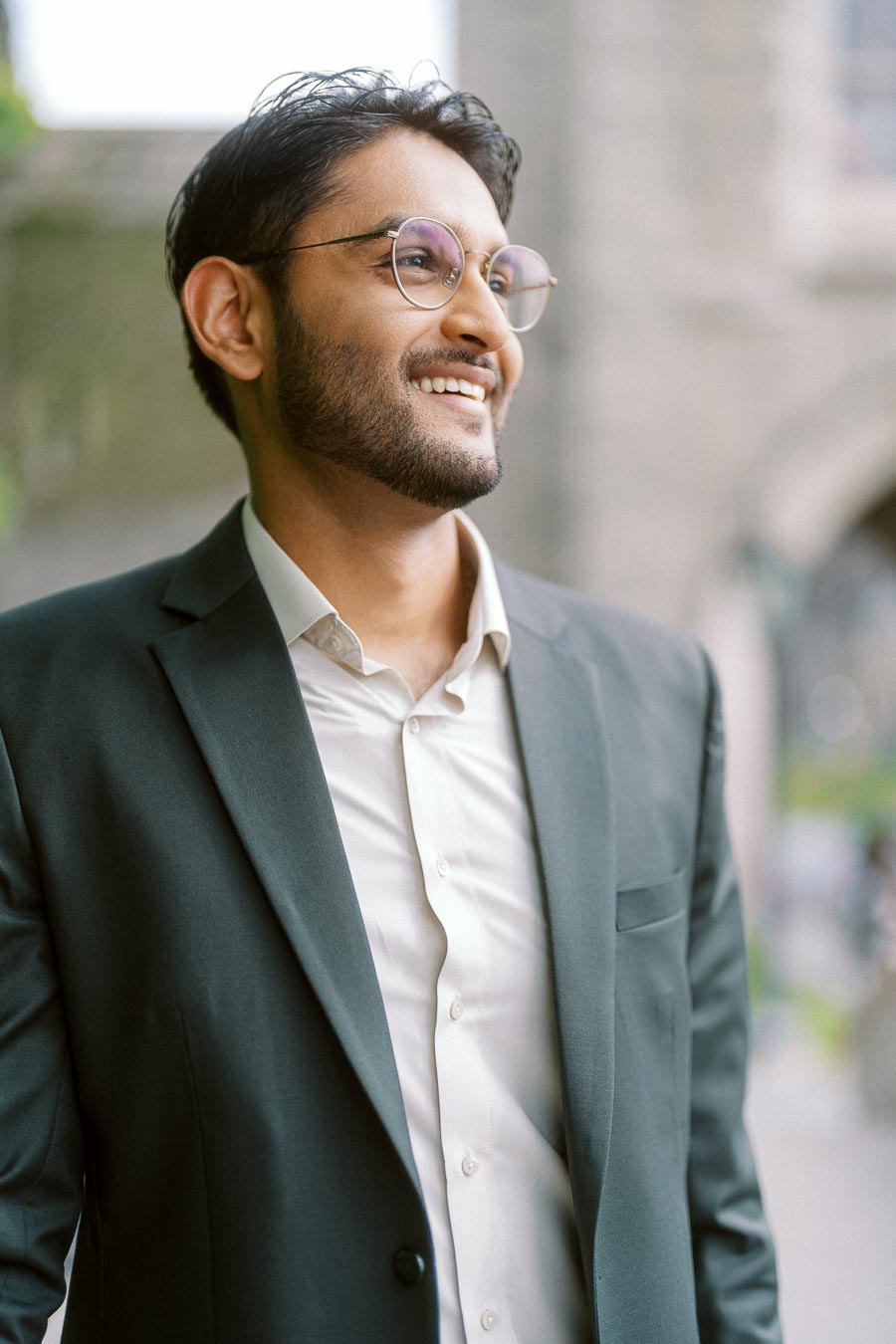 A man in a dark suit and glasses smiles while standing outdoors at the University of Toronto St. George campus, with blurred stone architecture in the background.